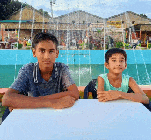 two young boys sitting at a table in front of a fountain with a sign that says ' shree ram '