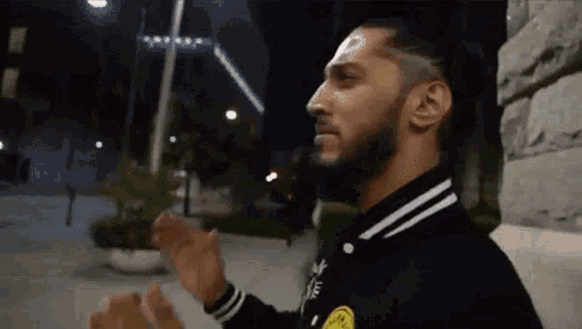 a man with a beard wearing a black adidas jacket is standing on a sidewalk at night .