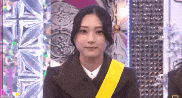 a woman wearing a black coat and a yellow sash is standing in front of a wall .