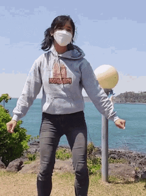 a woman wearing a mask and a hoodie with the word jerry on it is standing in front of the ocean .