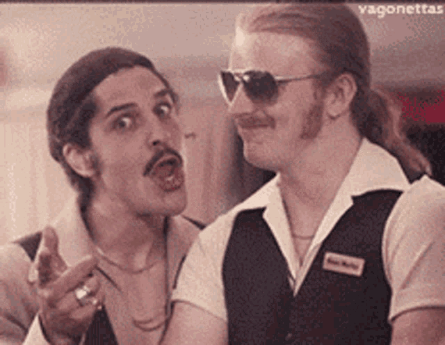 two men are standing next to each other and one of them is wearing sunglasses and a name tag .