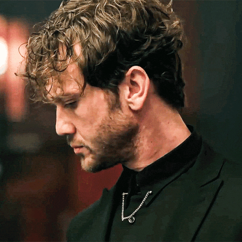 a man with curly hair and a beard wears a black suit and a necklace with the letter v on it