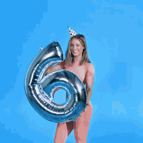 a woman wearing a party hat is holding a balloon in the shape of the number 6 .
