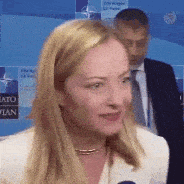 a woman in a white jacket is talking to a microphone while a man in a suit stands behind her .