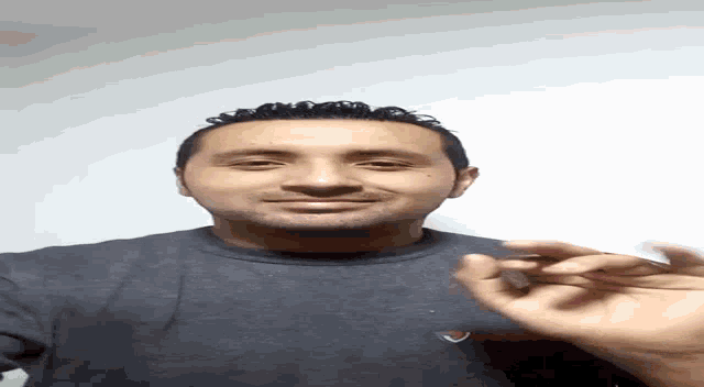 a man in a gray shirt is standing in front of a white wall with his hands in his pockets .
