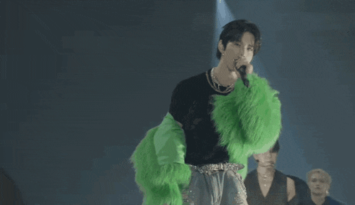 a man wearing a black shirt and a green fur coat singing into a microphone
