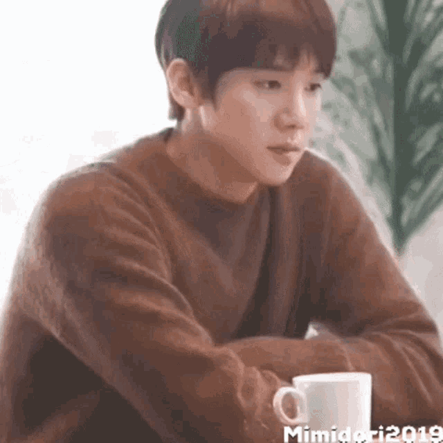 a young man in a brown sweater is sitting at a table holding a cup of coffee .