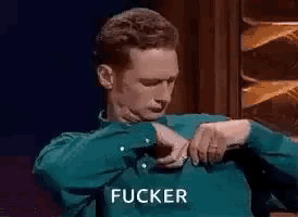 a man in a blue shirt is looking at his watch and saying `` fucker '' while sitting down .