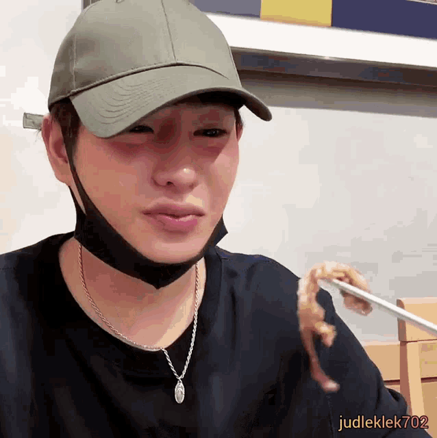 a man wearing a hat and a mask is eating something with chopsticks and the name judleklek702 is on the bottom