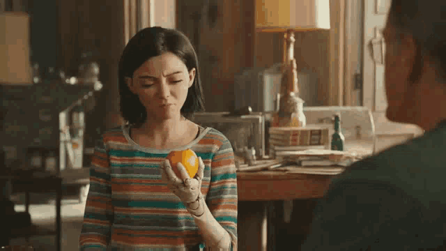 a woman with a prosthetic arm is holding an orange in her hand while a man points at her .