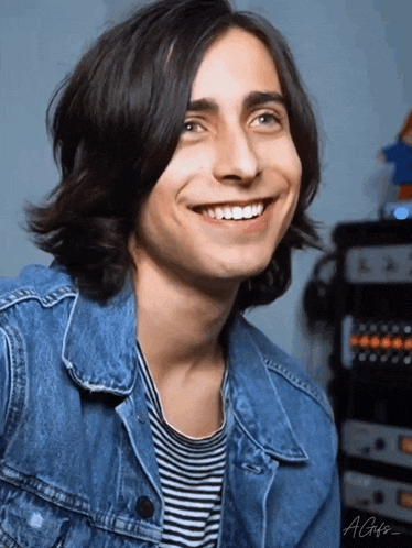a young man with long hair is smiling and wearing a denim jacket and striped shirt