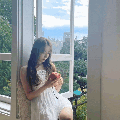 a woman in a white dress sits in front of a window