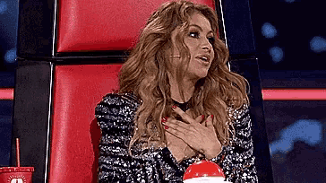 a woman in a sequined dress is sitting in a red chair holding her hands to her chest .