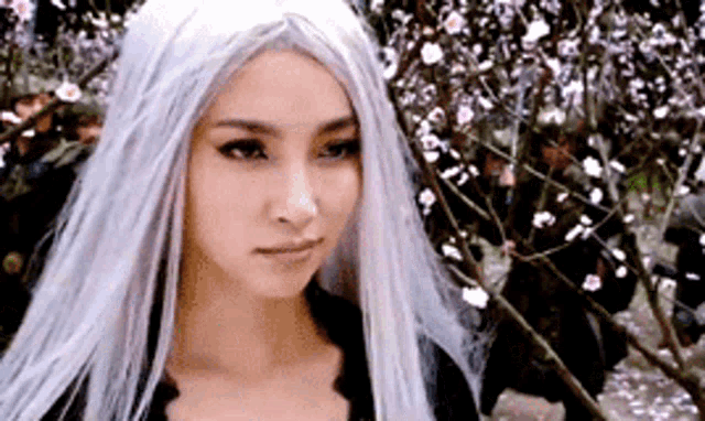 a woman with long white hair and a black dress is standing in front of a tree with flowers .
