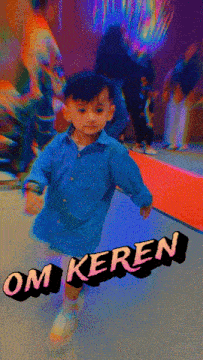 a little boy in a blue shirt is standing in front of a sign that says " om keren "