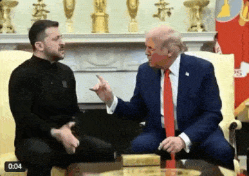 a man in a suit and tie is sitting next to a man in a black shirt and pointing at him .