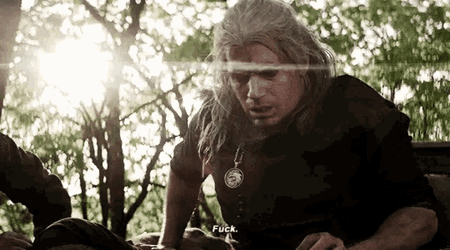 a man with long hair and a necklace is sitting in the woods and talking to another man .