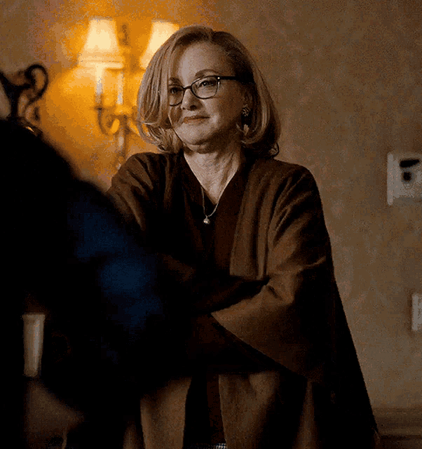 an older woman wearing glasses and a brown coat is standing in a room with her arms crossed .
