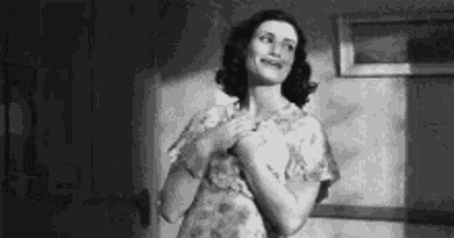 a black and white photo of a woman in a dress standing in a room with her hands on her chest .