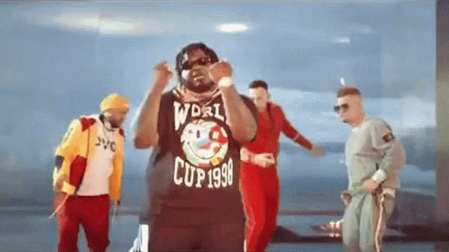 a group of men are dancing in a room with a man wearing a shirt that says world cup 1998 .