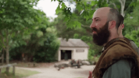 a bald man with a beard is standing in front of a house in a forest .