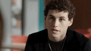 a young man with curly hair wearing a black jacket and a necklace
