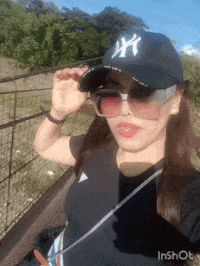 a woman wearing a ny hat and sunglasses is taking a selfie