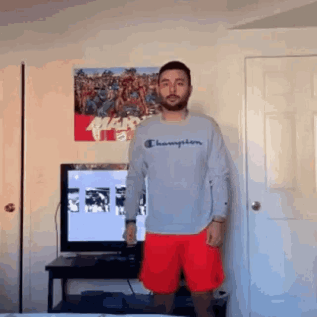 a man in a champion sweatshirt and red shorts is standing in front of a television in a living room .