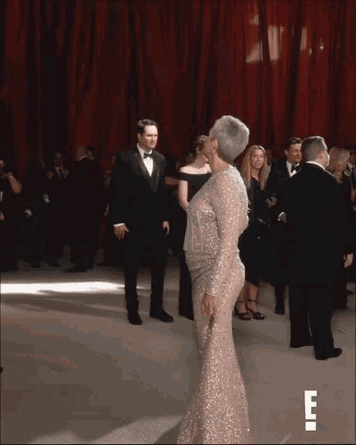 a woman in a sequined dress stands in front of a red curtain with the letter e on the bottom right