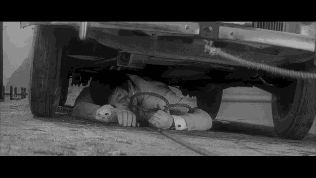 a black and white photo of a man laying under a car with a rope .