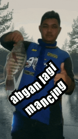 a man in a blue jacket holding a fish with the words " abyan lagi mancing " written on the bottom