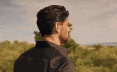 a man in a black shirt is standing in front of a field of trees and looking at the sky .