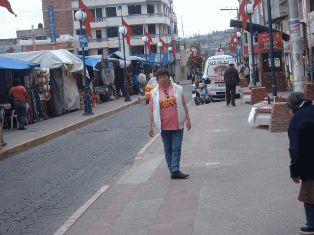 a woman in a pink shirt walks down a street with a white van that says ambulance