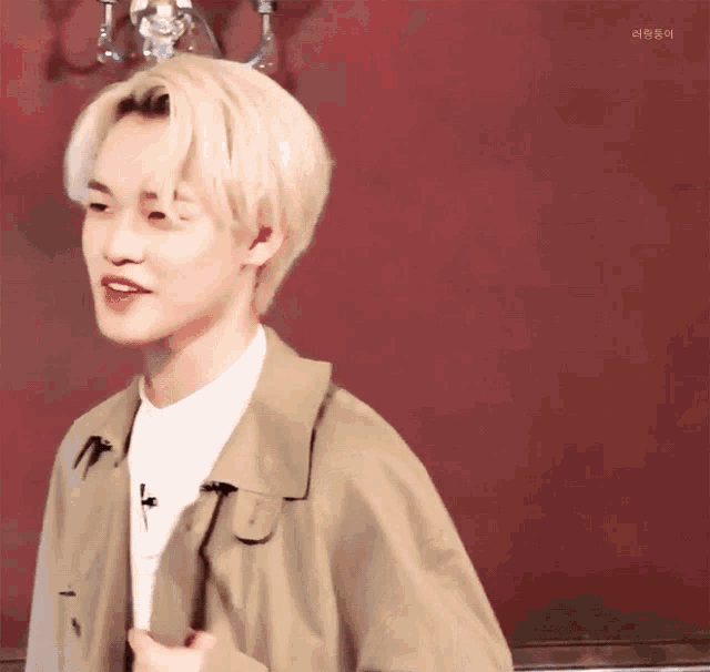 a young man with blonde hair and red lipstick is wearing a tan trench coat and a white shirt .