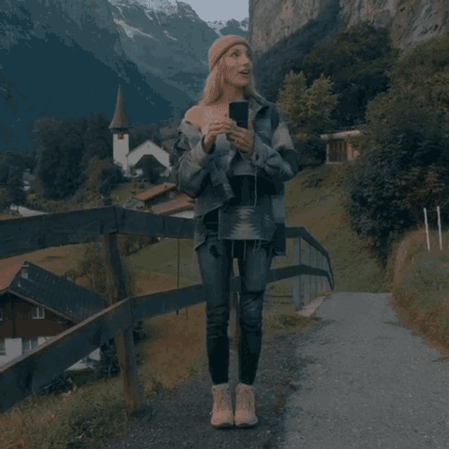a woman standing on a path holding a cell phone in front of a mountain