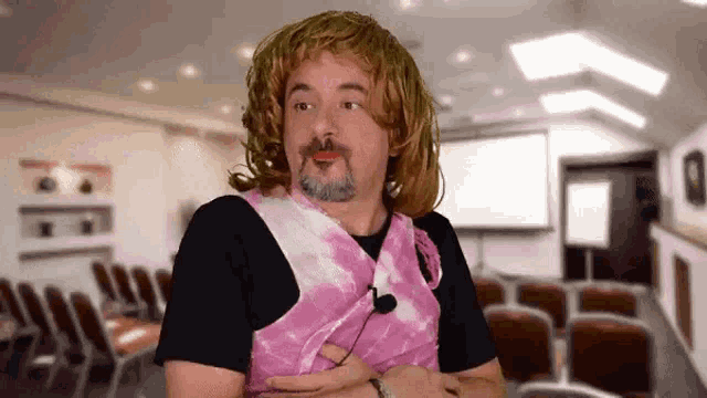 a man wearing a pink tie dye vest and a wig is standing in a conference room with his arms crossed .