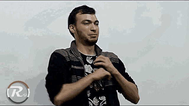 a man in a black jacket is making a gesture with his hands in front of a white background with the letter r on it