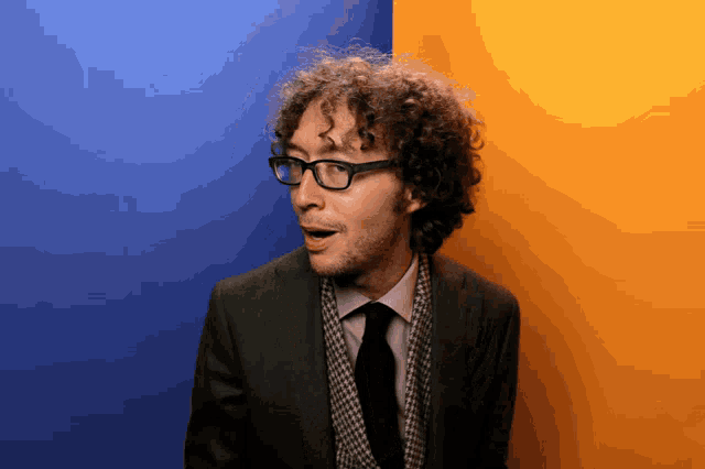 a man with curly hair wearing glasses and a suit stands in front of a blue and yellow wall