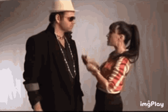 a man in a hat and sunglasses is talking to a woman in a red and white shirt .