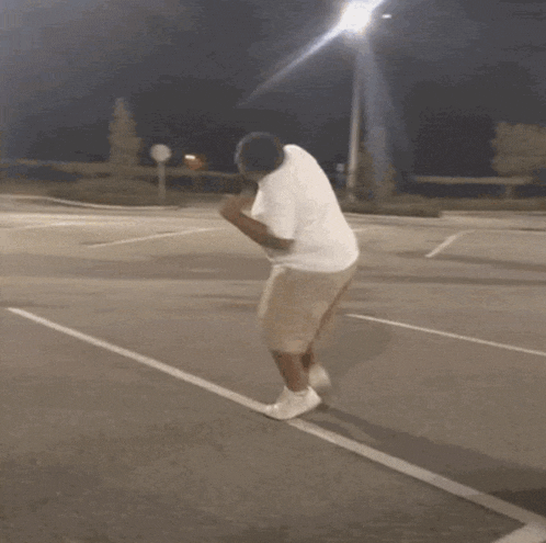 a man in a white shirt and shorts is dancing in a parking lot at night