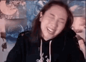 a woman in a black hoodie is laughing with her eyes closed while sitting in front of a computer screen .