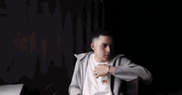 a young man wearing a gray hoodie and a white shirt is sitting at a table in front of a black curtain .