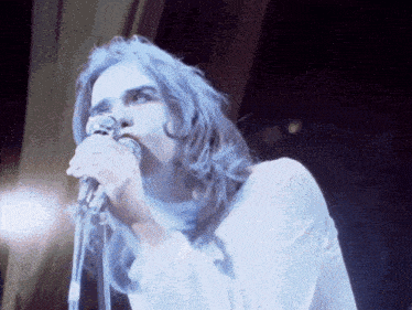 a man with long blue hair singing into a microphone while wearing a white shirt