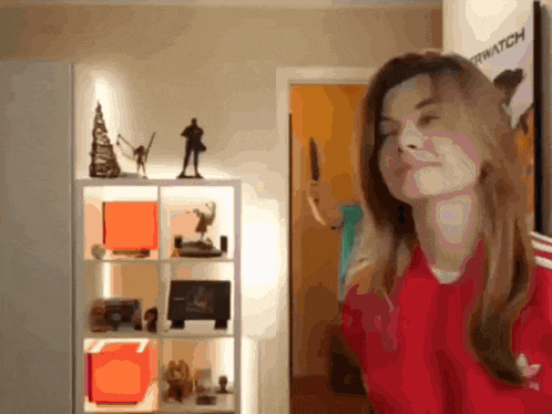 a woman in a red adidas shirt stands in front of a shelf with a poster that says overwatch