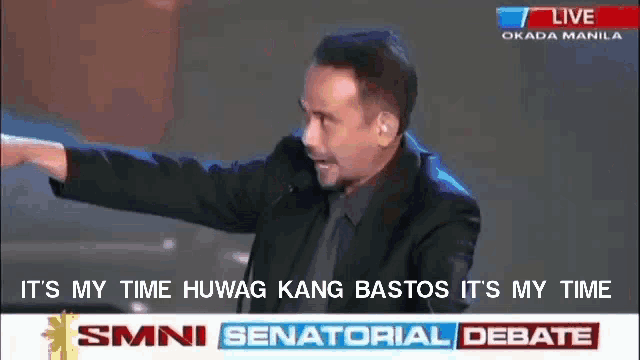 a man in a suit is giving a speech in front of a screen that says smni senatoral debate