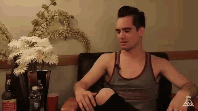 a man in a tank top sits in a chair with a vase of flowers in the background