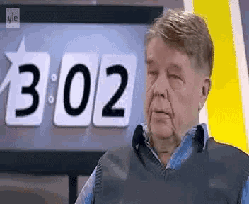 an older man is standing in front of a clock that says 3:02