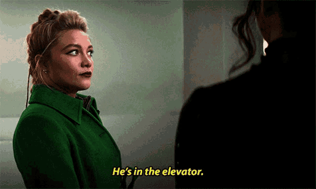 a woman in a green coat is talking to another woman in a dark room and says he 's in the elevator