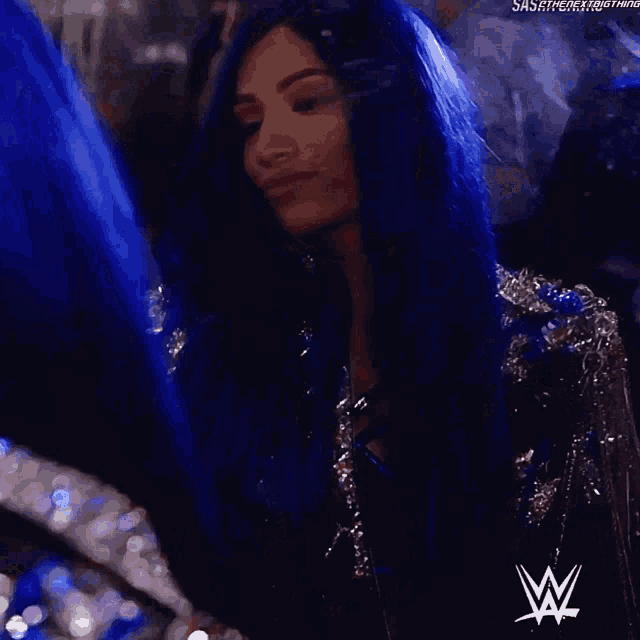 a woman with long blue hair is standing in a dark room with a w logo on her chest .