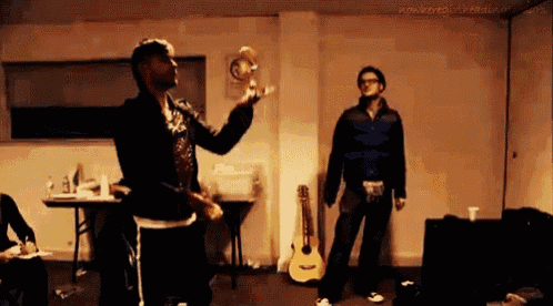 two men are standing in a room with a guitar in the background and a man holding a bottle in the foreground